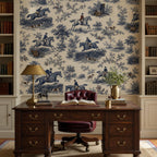 Cobalt blue equestrian toile wallpaper behind a carved wooden desk with a brass lamp and tufted burgundy chair